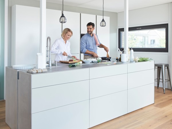Eine blonde Frau mit weißer Bluse und ein dunkelhaariger Mann in blauem Hemd kochen an einer Kochinsel