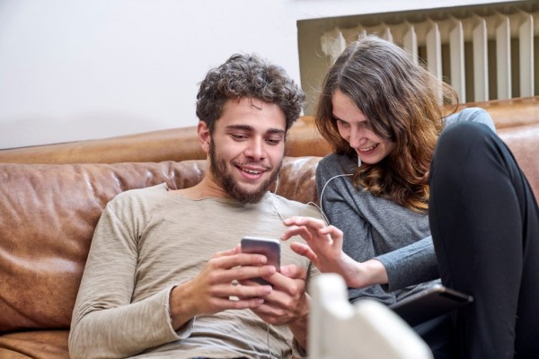 young man and young woman sitting on couch sharing cell phone and earphones