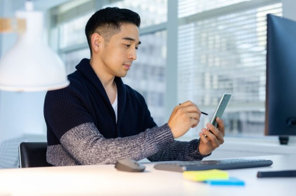 '一名男子坐在辦公桌前在Galaxy Note20上使用S筆。他面前是一台電腦顯示器，桌子上有鼠標，鍵盤和一些記事本。在他身後的窗戶中可以看到一座城市。