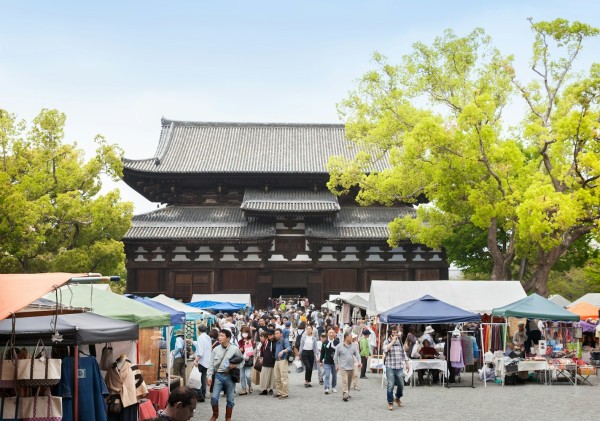 一大群人正在一个位于传统日本寺庙前的户外市场浏览各种摊位。五彩缤纷的遮阳篷覆盖着摊位，郁郁葱葱的树木在清澈的蓝天映衬下框住了这一场景。