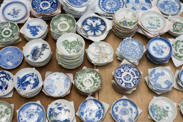 一堆各种各样的陶瓷盘子，展示着复杂的蓝色、绿色和白色图案，摆放在木制表面上。许多盘子上附有小红色价格标签。设计包括传统陶艺中常见的花卉、几何和动物图案。