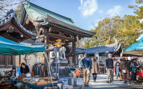 在一座传统日本寺庙前熙熙攘攘的户外市场场景。摊位上有各种物品，如古董和陶器。人们在蓝天之下浏览和聚集。寺庙的塔形屋顶在背景中可见，周围有树木。