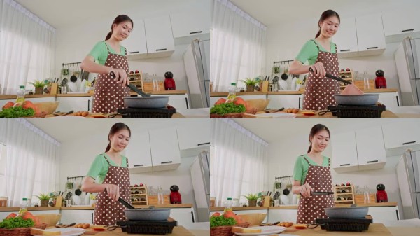 亚洲年轻女性在家中早晨的厨房里烹饪健康食品。迷人的女孩穿着围裙，感到快乐和放松，享受着使用平底锅准备