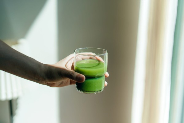 cropped hand of woman holding a glass of fresh green fruit and vegetable smoothie against white background with natural sunlight. healthy superfood. green colour, detox diet and healthy eating concept