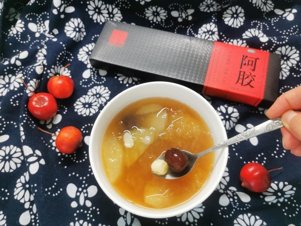 一位中年女性正在用研钵研磨西洋参片，旁边放置阿胶粉瓶和量勺，桌上铺有中式桌布，环境温馨整洁