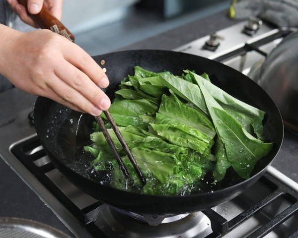 一位厨师正在用铁锅大火翻炒混合蔬菜，锅中腾起淡淡白烟，蔬菜呈现鲜绿色