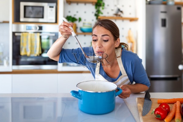 围着围裙的专注女性，用勺子品尝蓝色锅里的热汤。微笑的女厨师正在厨房检查自制餐点的味道。健康烹饪、居家生活和美食爱好概念。照片摄影图片