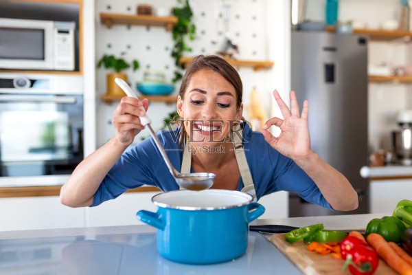 穿着围裙的快乐女子在厨房里,一边用勺子品尝蓝色锅里的汤,一边竖起大拇指。微笑的女厨师正在享用自制的餐点。健康烹饪、居家生活和成功的概念。照片摄影图片