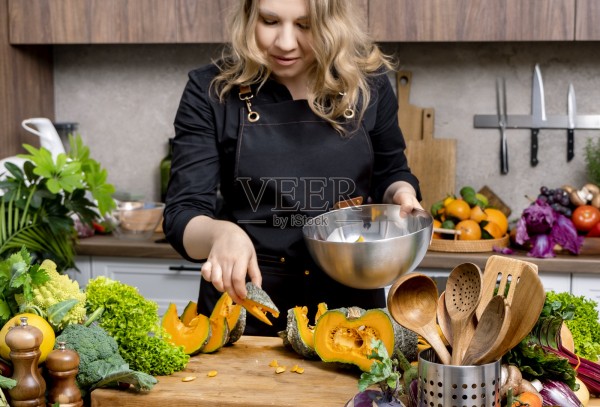 家庭厨师在明亮舒适的厨房里准备切好的南瓜,为健康的秋季餐做准备,周围有新鲜蔬菜和厨房工具。健康饮食氛围,烹饪生活方式概念。照片摄影图片